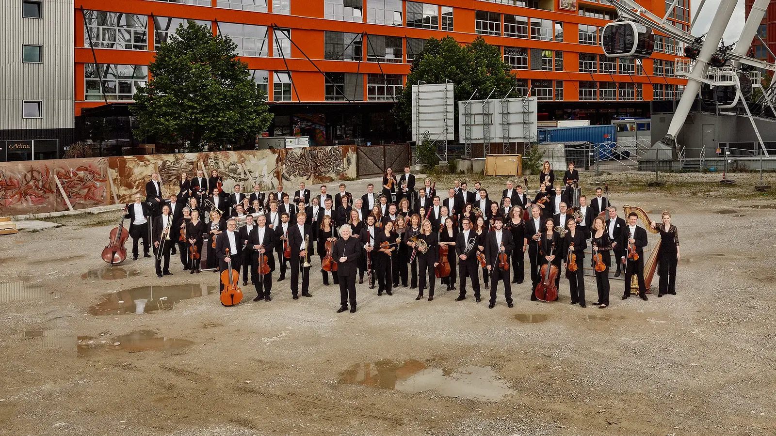 Das Orchester des Bayerischen Rundfunks, schwarz gekleidet, steht im freien auf einer ungeteerten Fläche mit Pfützen, im Hintergrund die Fassade eines großen Blocks