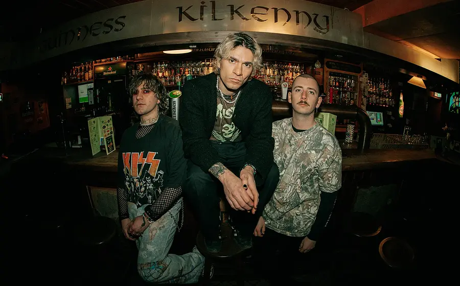Band‑Promofoto der Band TALCO in einer Pub‑Bar: Drei Personen posieren vor einer dicht bestückten Getränketheke; eine Person sitzt erhöht auf einem Barhocker in der Mitte, zwei Personen stehen links und rechts daneben. Über der Bar ist der Schriftzug „Guinness Kilkenny“ sichtbar, im Hintergrund Zapfhähne und Flaschenregale. Gedämpfte, warme Beleuchtung und Weitwinkel‑Perspektive erzeugen eine Club‑ und Punk‑Rock‑Atmosphäre, passend zu einem Live‑Konzert von TALCO.