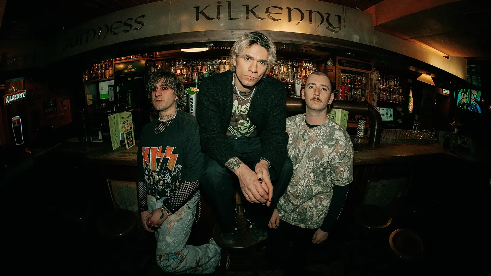 Band‑Promofoto der Band TALCO in einer Pub‑Bar: Drei Personen posieren vor einer dicht bestückten Getränketheke; eine Person sitzt erhöht auf einem Barhocker in der Mitte, zwei Personen stehen links und rechts daneben. Über der Bar ist der Schriftzug „Guinness Kilkenny“ sichtbar, im Hintergrund Zapfhähne und Flaschenregale. Gedämpfte, warme Beleuchtung und Weitwinkel‑Perspektive erzeugen eine Club‑ und Punk‑Rock‑Atmosphäre, passend zu einem Live‑Konzert von TALCO.