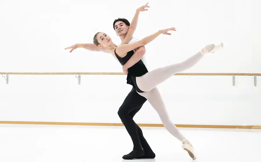 Ballettpaar in einem hellen Tanzsaal vor einer Ballettstange. Eine Person in Spitzenschuhen streckt ein Bein waagerecht aus, während die andere Person die Figur hebt und beide die Arme elegant ausbreiten.