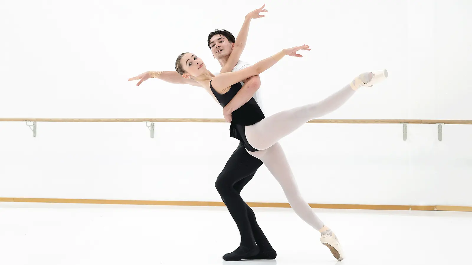 Ballettpaar in einem hellen Tanzsaal vor einer Ballettstange. Eine Person in Spitzenschuhen streckt ein Bein waagerecht aus, während die andere Person die Figur hebt und beide die Arme elegant ausbreiten.