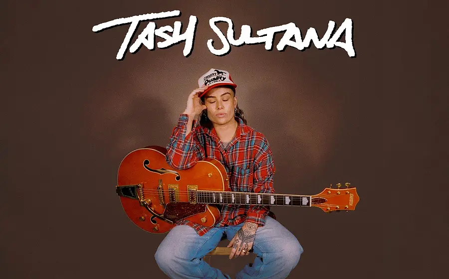 Eventfoto „TASH SULTANA“ mit der Künstlerin auf einem Hocker mit Saiteninstrument vor braunem Hintergrund.