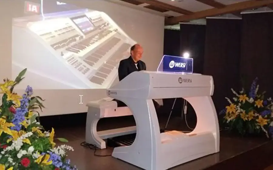 Heiko Schneider spielt an einer modernen weißen Orgel auf einer Bühne, im Hintergrund eine projizierte Orgelansicht und Blumenarrangements