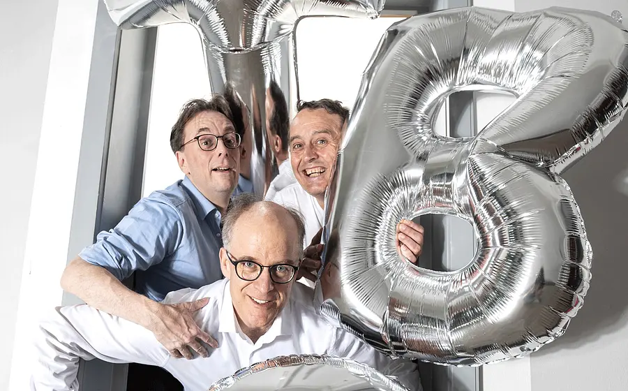 Drei Personen stehen in einem Türrahmen zwischen silbernen Folienballons in Form der Buchstaben T B und C und blicken in die Kamera