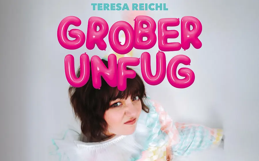 Die Künstlerin Theresa Reichl mit Grimasse und pinker Aufschrift Grober Unfug vor hellgrau verschwommenem Hintergrund