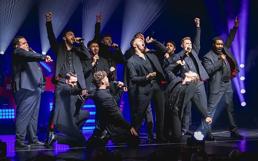 Zwölf Darsteller der 12 Tenors in dunklen Anzügen posieren auf einer Bühne in violett-blauem Licht
