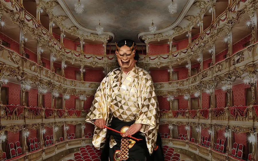 Person in einem reich verzierten gold‑schwarzen Noh‑Kostüm steht auf der Bühne des Cuvilliés‑Theaters; hinter der Person sind die ornamentreichen roten und goldenen Ränge und Balkone des historischen Theaters zu sehen