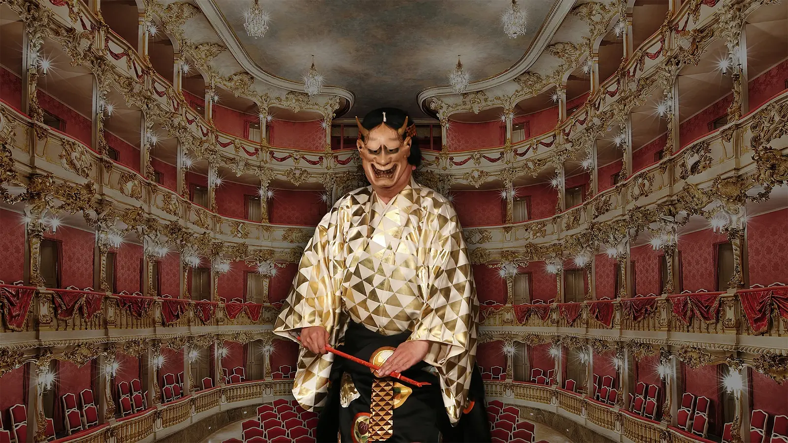 Person in einem reich verzierten gold‑schwarzen Noh‑Kostüm steht auf der Bühne des Cuvilliés‑Theaters; hinter der Person sind die ornamentreichen roten und goldenen Ränge und Balkone des historischen Theaters zu sehen