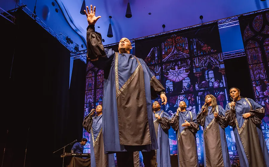 Bühnenfoto von Best of Black Gospel mit mehreren Sängerinnen und Sängern in blau grauen Chorgewändern auf einer Konzertbühne, eine Person steht mit erhobener Hand im Vordergrund, im Hintergrund stilisierte Kirchenfenster und Bühnenbeleuchtung