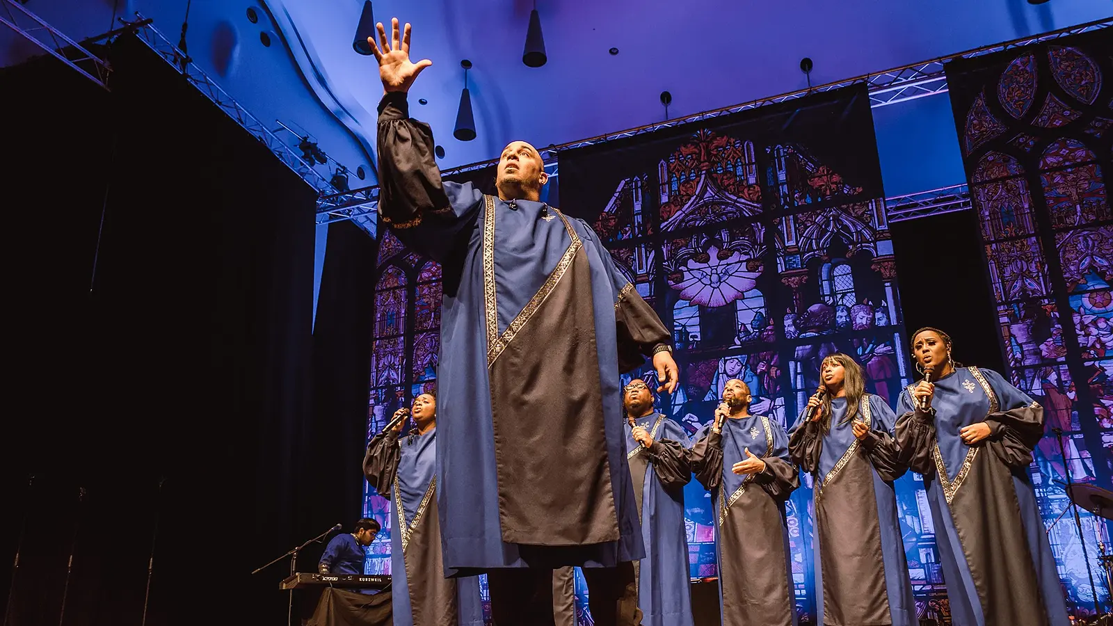 Bühnenfoto von Best of Black Gospel mit mehreren Sängerinnen und Sängern in blau grauen Chorgewändern auf einer Konzertbühne, eine Person steht mit erhobener Hand im Vordergrund, im Hintergrund stilisierte Kirchenfenster und Bühnenbeleuchtung