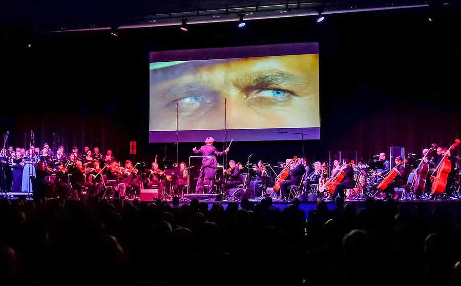 Bühnenfoto eines Orchesters mit Chor und Dirigent vor Publikum, darüber eine große Leinwand mit einer Nahaufnahme blauer Augen aus einer Filmszene, auf einer rot violett beleuchteten Konzertbühne zur Veranstaltung The Best of Ennio Morricone