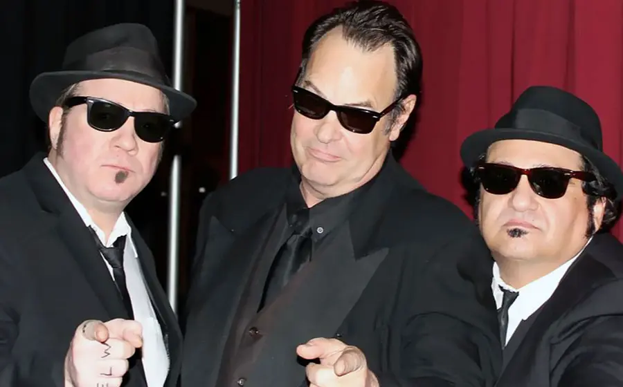Drei Männer in schwarzen Anzügen, Hüten und Sonnenbrillen im Blues-Brothers-Stil, die in die Kamera zeigen – Motiv zur Show „The Official Blues Brothers Revue“.