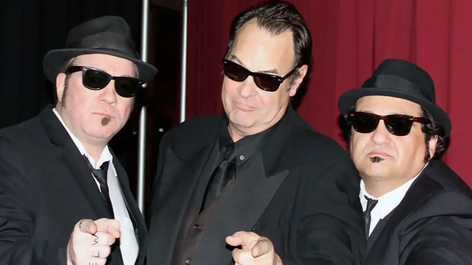Drei Männer in schwarzen Anzügen, Hüten und Sonnenbrillen im Blues-Brothers-Stil, die in die Kamera zeigen – Motiv zur Show „The Official Blues Brothers Revue“.