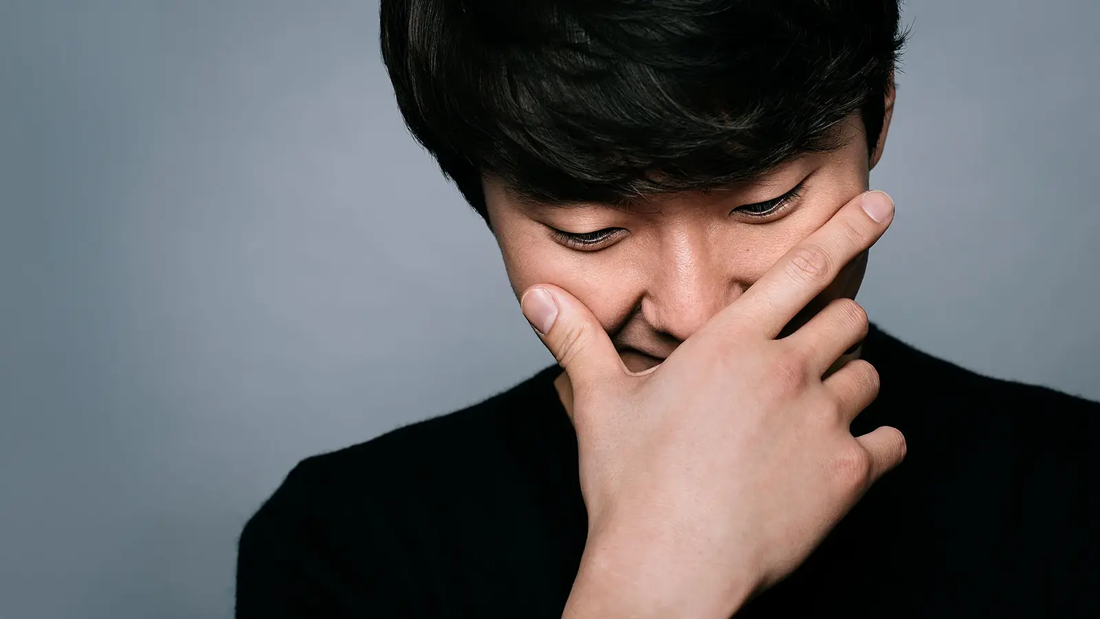 Porträtmotiv von Seong‑Jin Cho: Der Pianist ist in enger Nahaufnahme zu sehen und hält eine Hand vor das Gesicht, der Kopf ist leicht nach unten geneigt. Er trägt ein dunkles Oberteil. Neutraler grauer Studiohintergrund ohne weitere Elemente. Weiches, gerichtetes Licht betont Gesicht und Hand. Ruhiges, konzentriertes Promofoto für einen klassischen Klavierabend mit Seong‑Jin Cho.