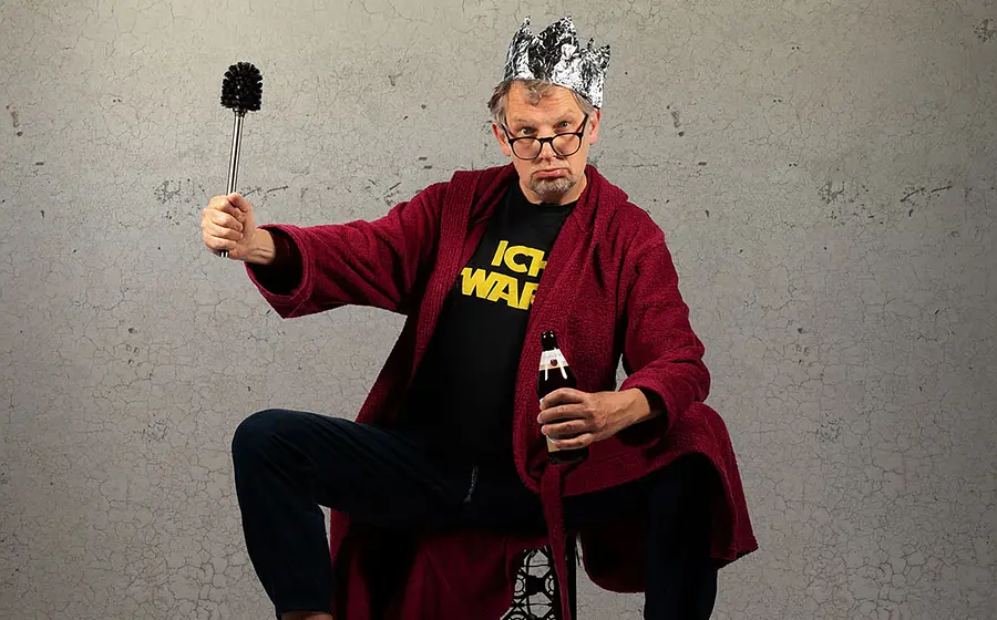 Foto für die Veranstaltung von Severin Groebner – ICH BIN DAS VOLK mit dem Künstler, der auf einem Stuhl sitzt, in einer Hand ein Bier hält und in der anderen eine Toilettenbürste, während er eine Krone aus Alufolie trägt; im Hintergrund eine Steinwand
