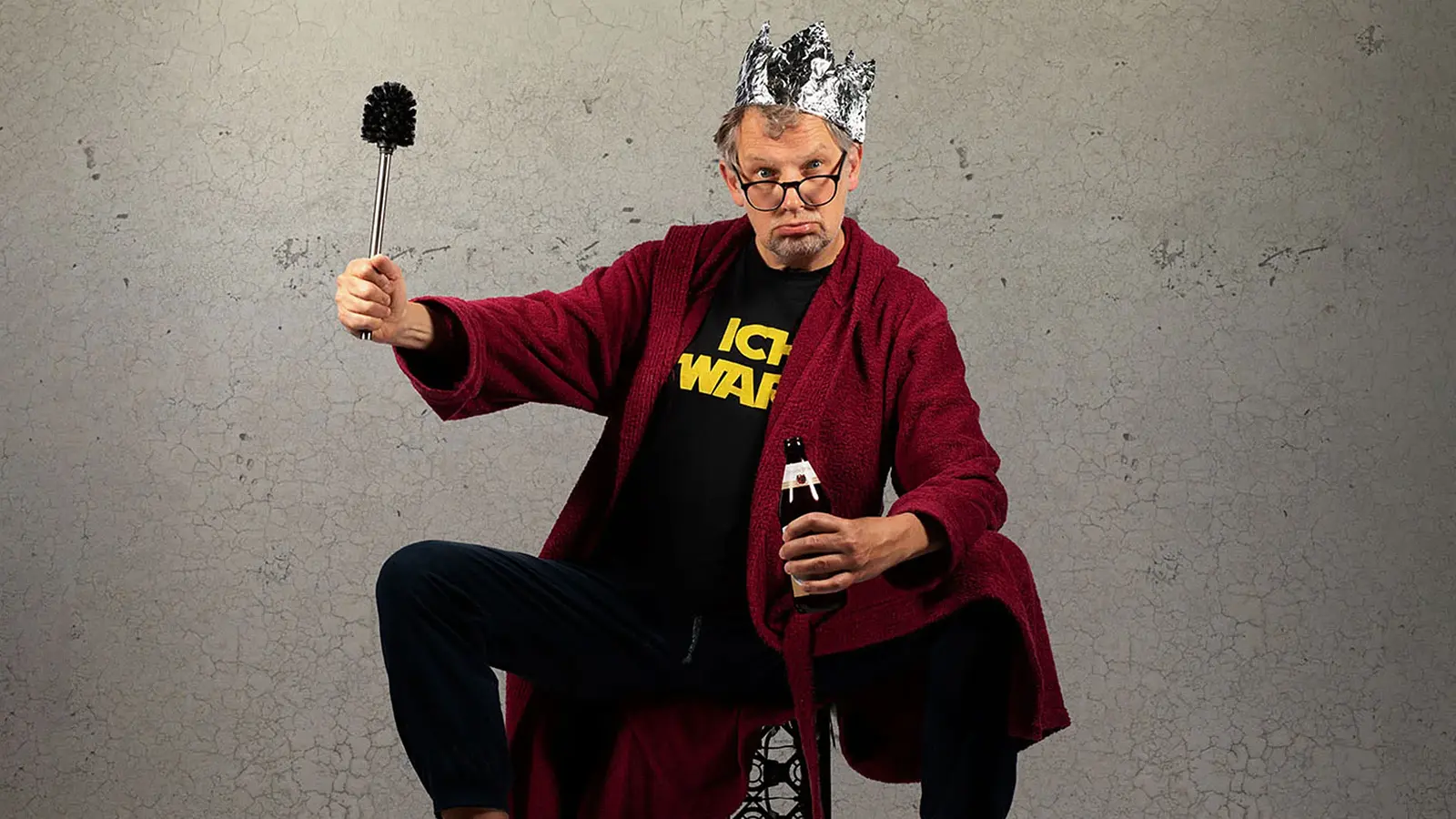 Foto für die Veranstaltung von Severin Groebner – ICH BIN DAS VOLK mit dem Künstler, der auf einem Stuhl sitzt, in einer Hand ein Bier hält und in der anderen eine Toilettenbürste, während er eine Krone aus Alufolie trägt; im Hintergrund eine Steinwand