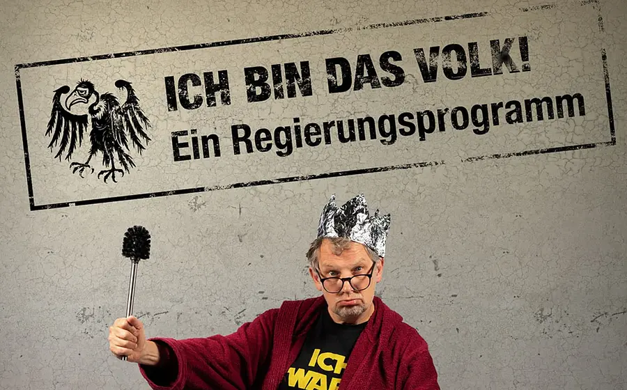 Plakatmotiv mit den Aufschriften Ich bin das Volk! und Ein Regierungsprogramm. Eine Person mit Krone aus Alufolie, Brille und rotem Bademantel hält eine Toilettenbürste wie ein Zepter vor grauem Hintergrund.
