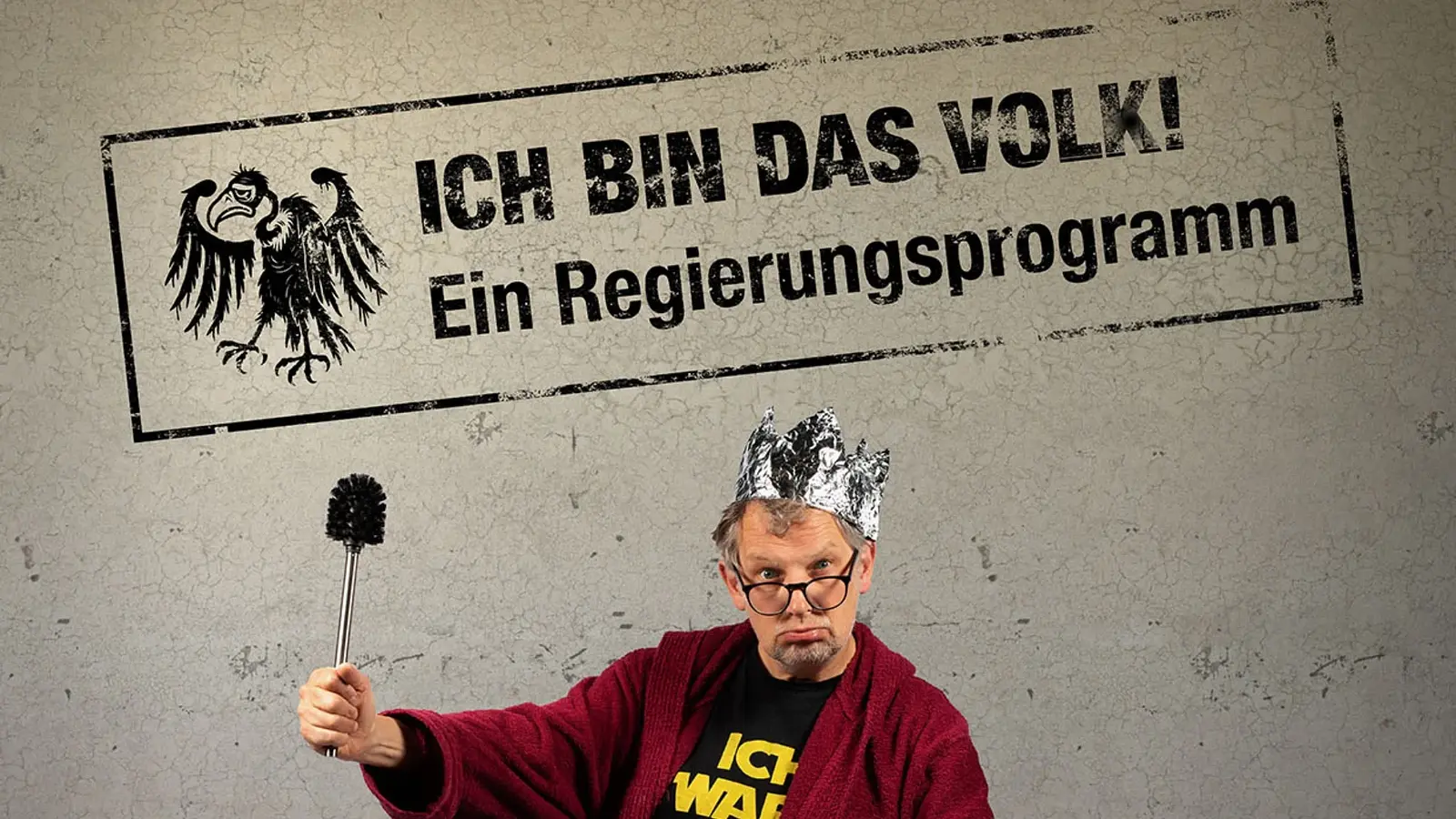 Plakatmotiv mit den Aufschriften Ich bin das Volk! und Ein Regierungsprogramm. Eine Person mit Krone aus Alufolie, Brille und rotem Bademantel hält eine Toilettenbürste wie ein Zepter vor grauem Hintergrund.