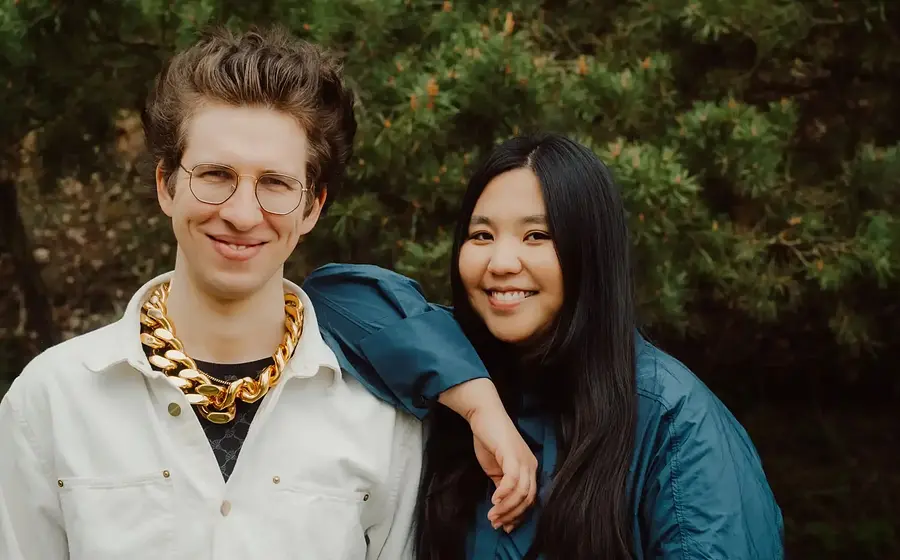 Shuteen Erdenebaatar und Nils Kugelmann vor dichtem Grün, er mit runder Brille, hellem Hemd und auffälliger goldfarbener Kette, sie in dunkelblauer Jacke mit langem Haar, beide nebeneinander in die Kamera blickend.