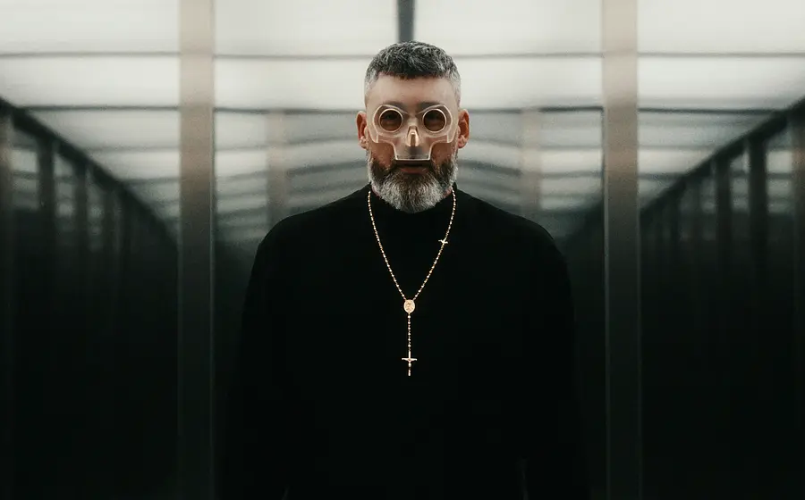 Sido mit transparenter Maske und Kreuzkette steht in einem symmetrischen, spiegelnden Gang mit futuristischer Atmosphäre.