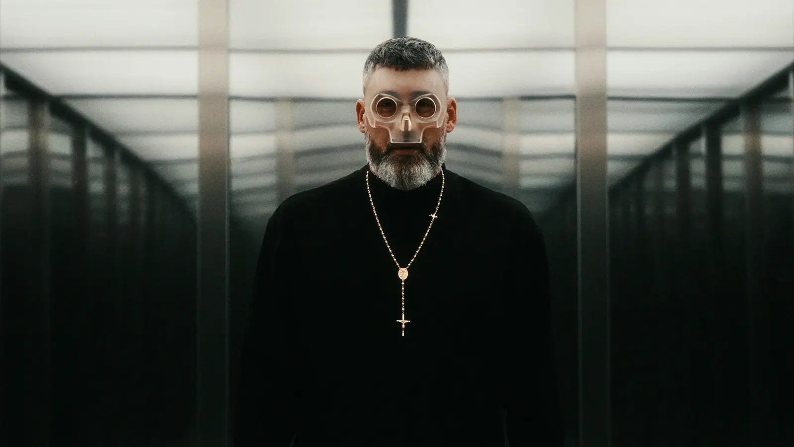 Sido mit transparenter Maske und Kreuzkette steht in einem symmetrischen, spiegelnden Gang mit futuristischer Atmosphäre.
