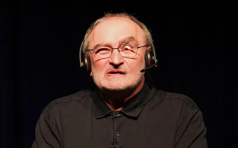 Portrait von Sigi Zimmerschied, der eine Grimasse macht, in schwarzem Hemd, mit Kopfhörern, vor schwarzem Hintergrund