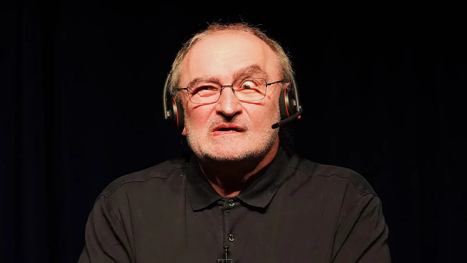 Portrait von Sigi Zimmerschied, der eine Grimasse macht, in schwarzem Hemd, mit Kopfhörern, vor schwarzem Hintergrund