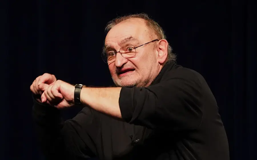 Sigi Zimmerschied mit Brille und schwarzem Hemd steht auf einer Bühne vor dunklem Hintergrund und zeigt mit gestikulierenden Armen eine ausdrucksstarke Geste während eines Kabarettauftritts.