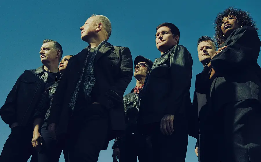 Die sieben Bandmitglieder von Simple Mind in dunkler Kleidung stehen im Freien vor blauem Himmel. Die Aufnahme wurde aus einer niedrigen Perspektive fotografiert.