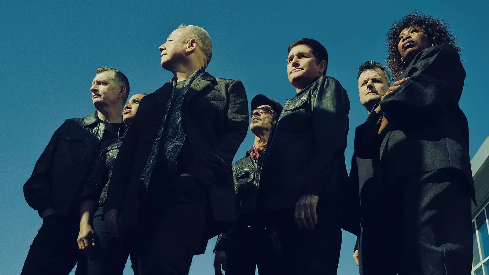 Die sieben Bandmitglieder von Simple Mind in dunkler Kleidung stehen im Freien vor blauem Himmel. Die Aufnahme wurde aus einer niedrigen Perspektive fotografiert.