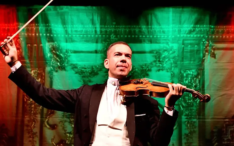 Person in festlicher Konzertkleidung hält eine Violine und einen erhobenen Geigenbogen auf einer Bühne. Im Hintergrund rot-grüne Beleuchtung und ein dekorativer Bühnenvorhang.