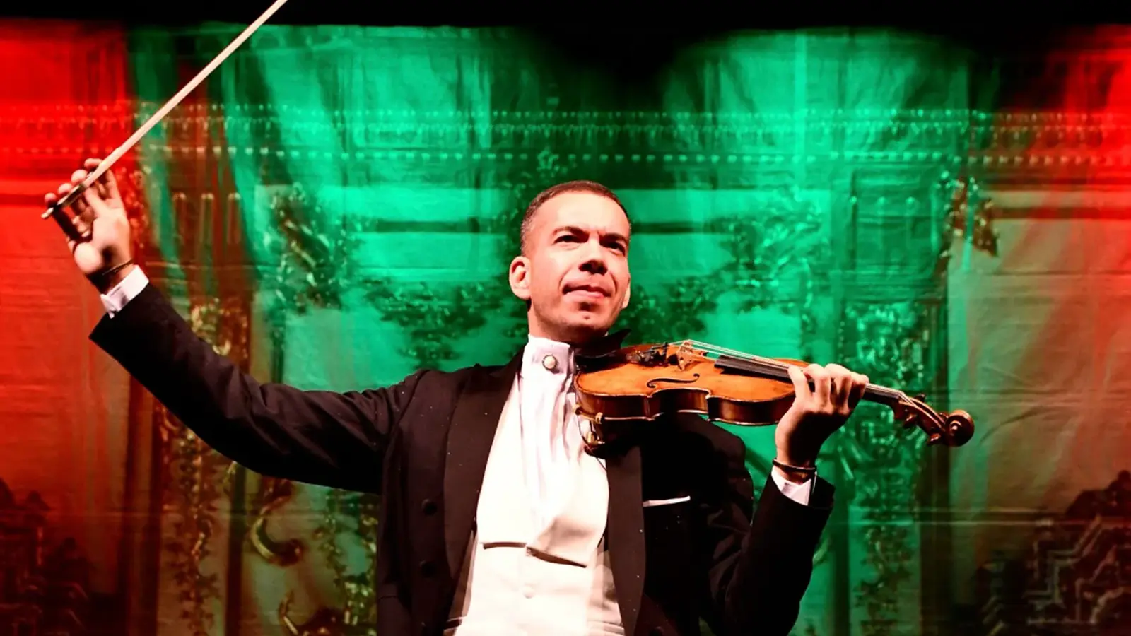 Person in festlicher Konzertkleidung hält eine Violine und einen erhobenen Geigenbogen auf einer Bühne. Im Hintergrund rot-grüne Beleuchtung und ein dekorativer Bühnenvorhang.