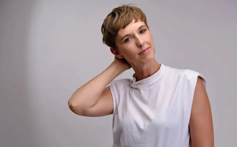 Sonja Pikart in weißem ärmellosem Oberteil vor hellem grauem Hintergrund, mit kurzem Haar, zur Seite geneigtem Kopf und einer Hand im Nacken.