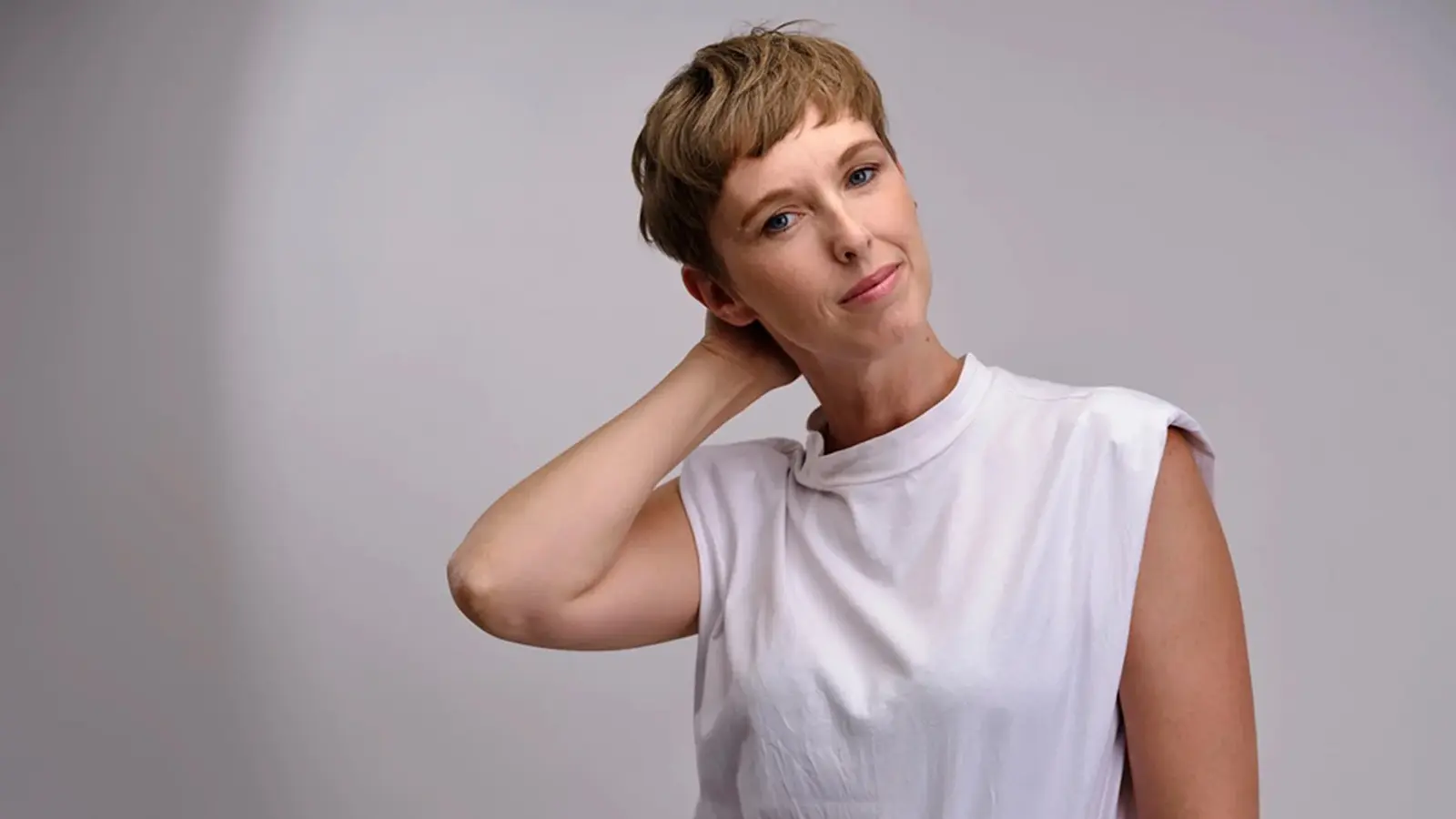 Sonja Pikart in weißem ärmellosem Oberteil vor hellem grauem Hintergrund, mit kurzem Haar, zur Seite geneigtem Kopf und einer Hand im Nacken.