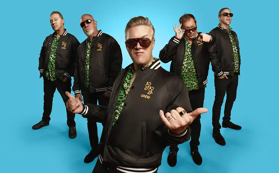 Studiofoto von fünf Bandmitgliedern der Spike and the Gimme Gimmes in schwarzen Jacken und grün gemusterten Hemden vor blauem Hintergrund, eine Person im Vordergrund mit Sonnenbrille und Handgeste