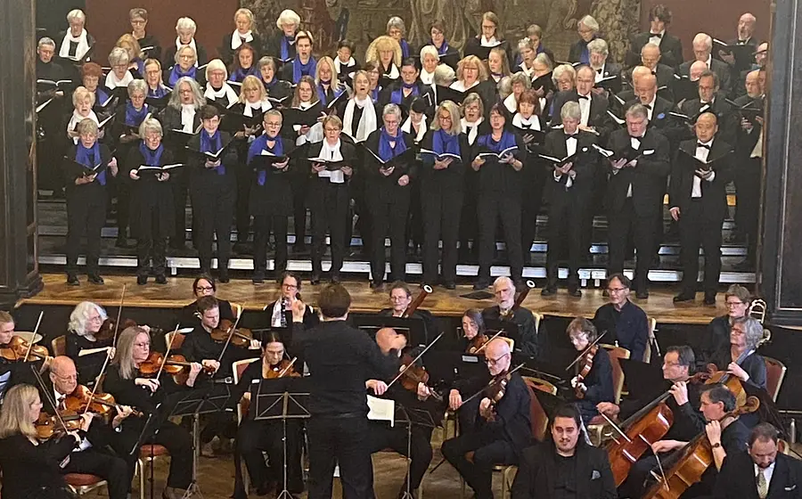 Konzertaufführung mit großem Chor auf gestuften Podesten und Sinfonieorchester im Vordergrund. Die Mitwirkenden tragen schwarze Konzertkleidung mit weißen und blauen Schals bzw. Fliegen; in der Mitte dirigiert eine Person das Ensemble