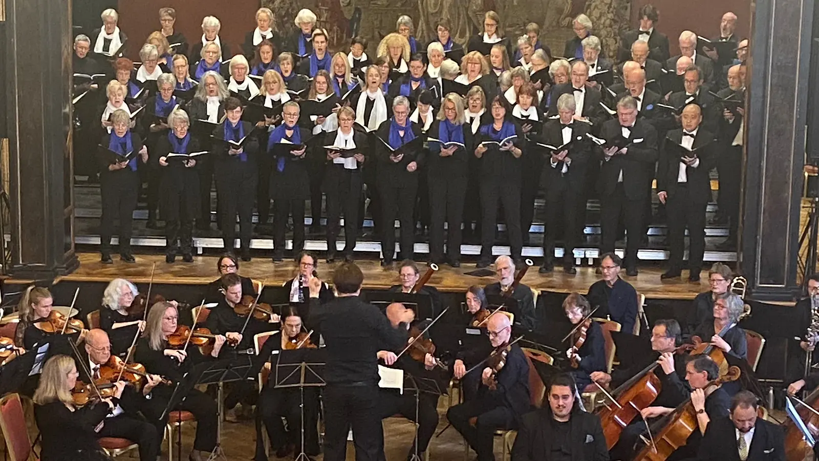 Konzertaufführung mit großem Chor auf gestuften Podesten und Sinfonieorchester im Vordergrund. Die Mitwirkenden tragen schwarze Konzertkleidung mit weißen und blauen Schals bzw. Fliegen; in der Mitte dirigiert eine Person das Ensemble