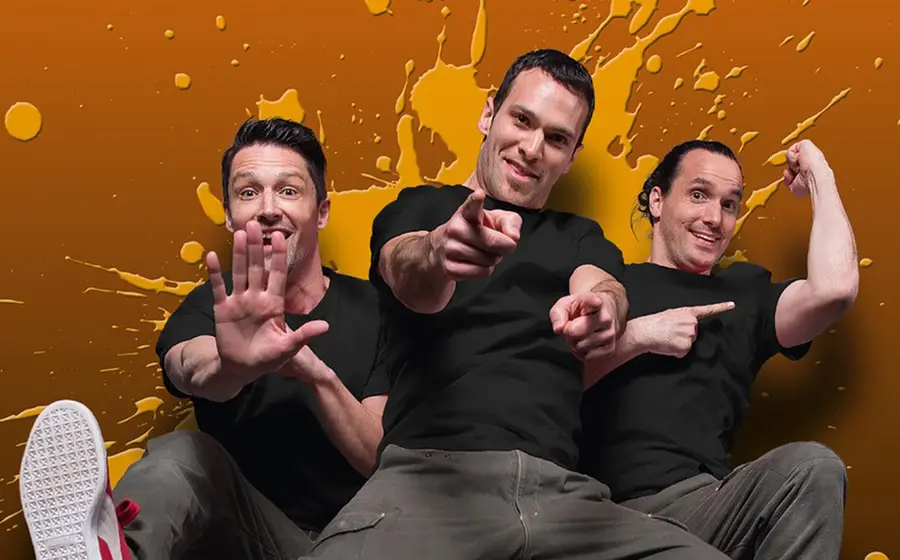 Foto zu „Starbugs Comedy – JUMP! Reloaded“ mit drei Männern in schwarzen T-Shirts vor orangefarbenem Hintergrund, in dynamischen Posen.