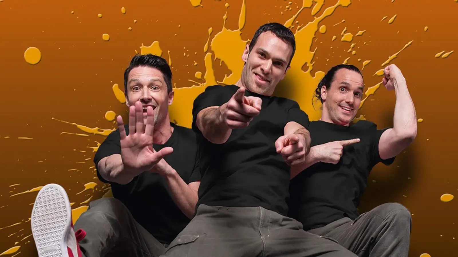 Foto zu „Starbugs Comedy – JUMP! Reloaded“ mit drei Männern in schwarzen T-Shirts vor orangefarbenem Hintergrund, in dynamischen Posen.