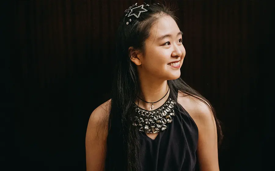 Tianyao Lyu mit langen dunklen Haaren vor dunklem Hintergrund, trägt ein schwarzes Oberteil mit auffälligem Schmuck am Ausschnitt und blickt seitlich.