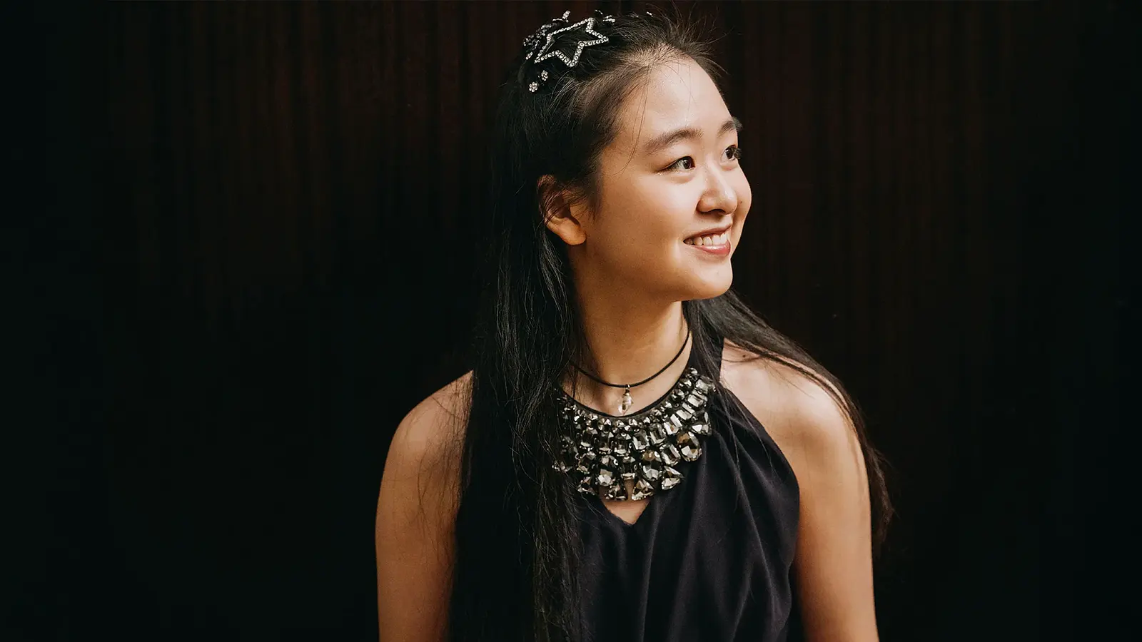 Tianyao Lyu mit langen dunklen Haaren vor dunklem Hintergrund, trägt ein schwarzes Oberteil mit auffälligem Schmuck am Ausschnitt und blickt seitlich.