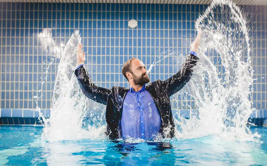 Tobi Freudenthal steht in Hemd und Sakko bis zur Brust in einem Hallenbad und schleudert mit erhobenen Armen Wasserfontänen durch die Luft vor blau gefliester Wand