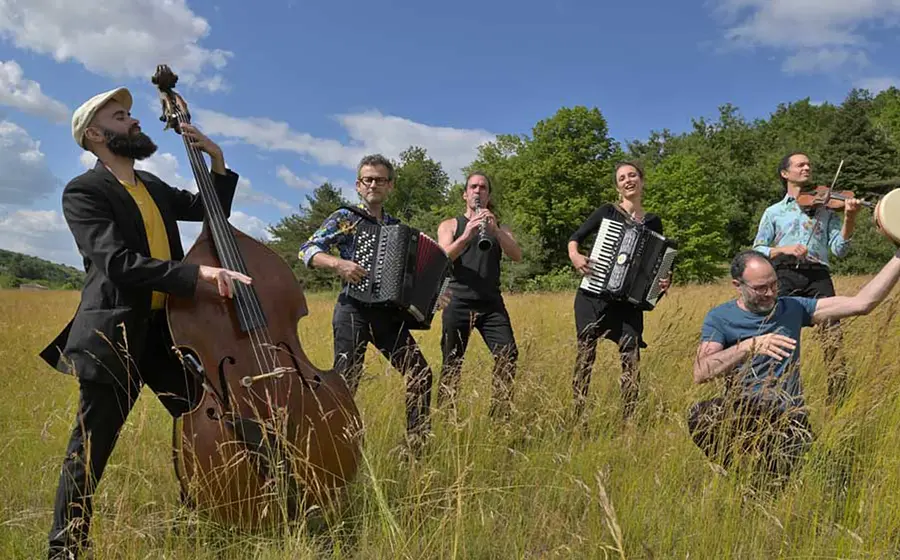 Bandfoto von Tram des Balkans mit sechs Musikerinnen und Musikern auf einer Wiese, darunter Kontrabass, Akkordeons, Klarinette, Violine und Rahmentrommel, vor Bäumen und blauem Himmel