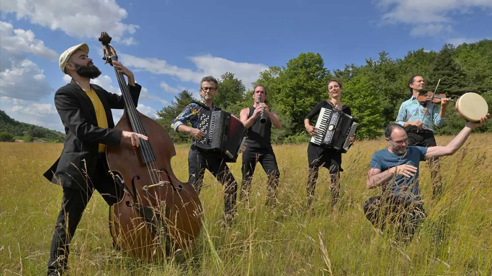 Bandfoto von Tram des Balkans mit sechs Musikerinnen und Musikern auf einer Wiese, darunter Kontrabass, Akkordeons, Klarinette, Violine und Rahmentrommel, vor Bäumen und blauem Himmel