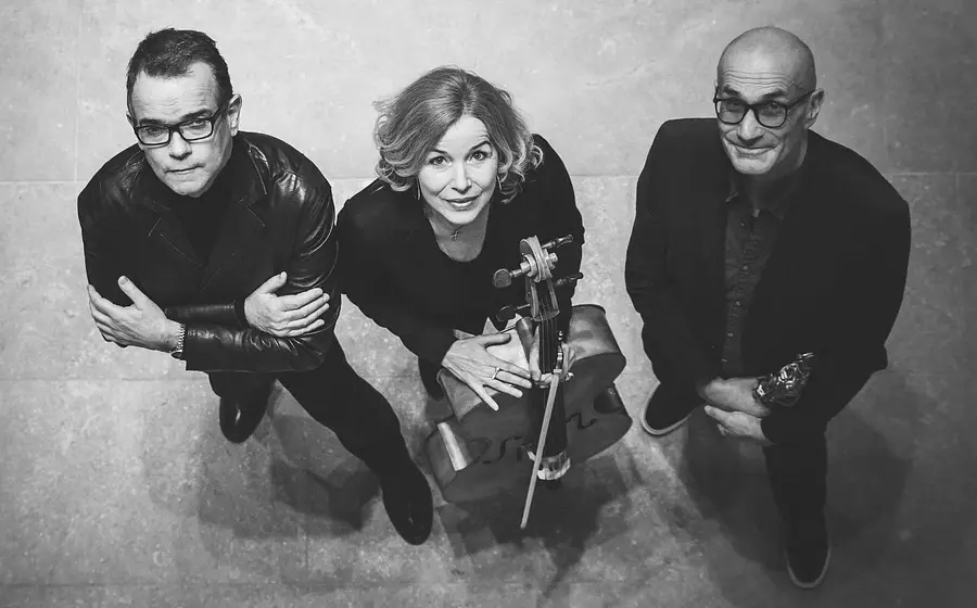 Schwarzweißes Pressefoto des Trio Sfera mit drei Musikerinnen und Musikern von oben fotografiert, eine Person mit Violoncello in der Mitte, flankiert von zwei Personen in dunkler Kleidung, vor hellem Steinboden.