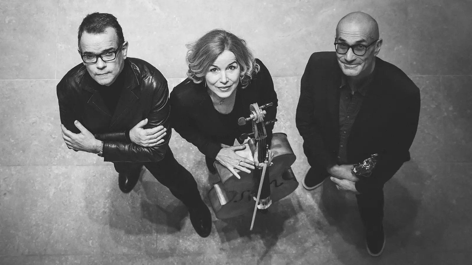 Schwarzweißes Pressefoto des Trio Sfera mit drei Musikerinnen und Musikern von oben fotografiert, eine Person mit Violoncello in der Mitte, flankiert von zwei Personen in dunkler Kleidung, vor hellem Steinboden.
