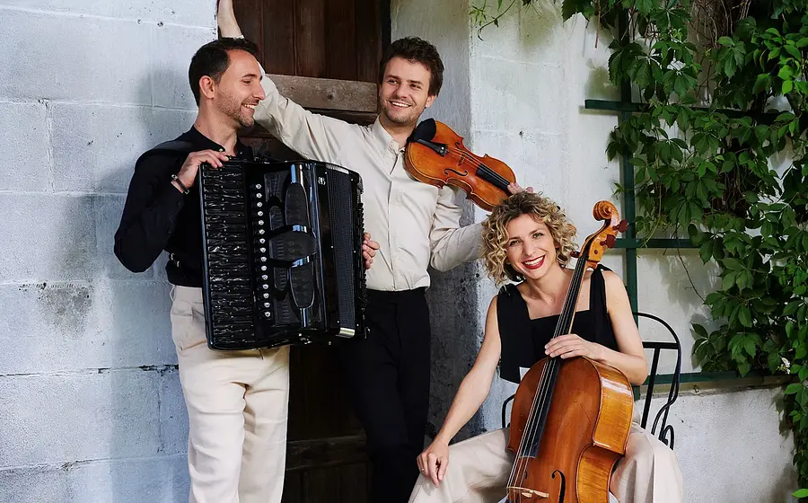 Bandfoto Trio Wellcaru mit drei Personen vor einer weßen Ziegelwand mit Holztür, eine Person mit Akkordeon eine mit Geige und eine mit Cello daneben rankende Pflanzen