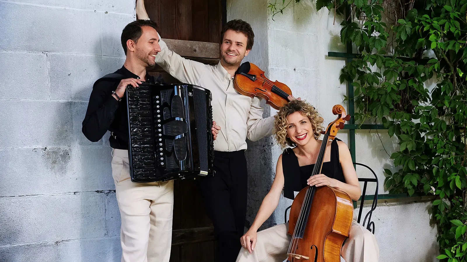 Bandfoto Trio Wellcaru mit drei Personen vor einer weßen Ziegelwand mit Holztür, eine Person mit Akkordeon eine mit Geige und eine mit Cello daneben rankende Pflanzen