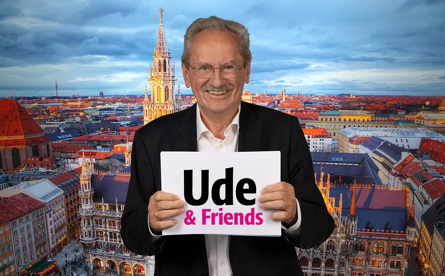 Person mit Brille hält ein Schild mit der Aufschrift Ude & Friends vor sich, im Hintergrund Panorama der Münchner Altstadt mit Rathaus, Kirchturm und Dächern bei Abendlicht