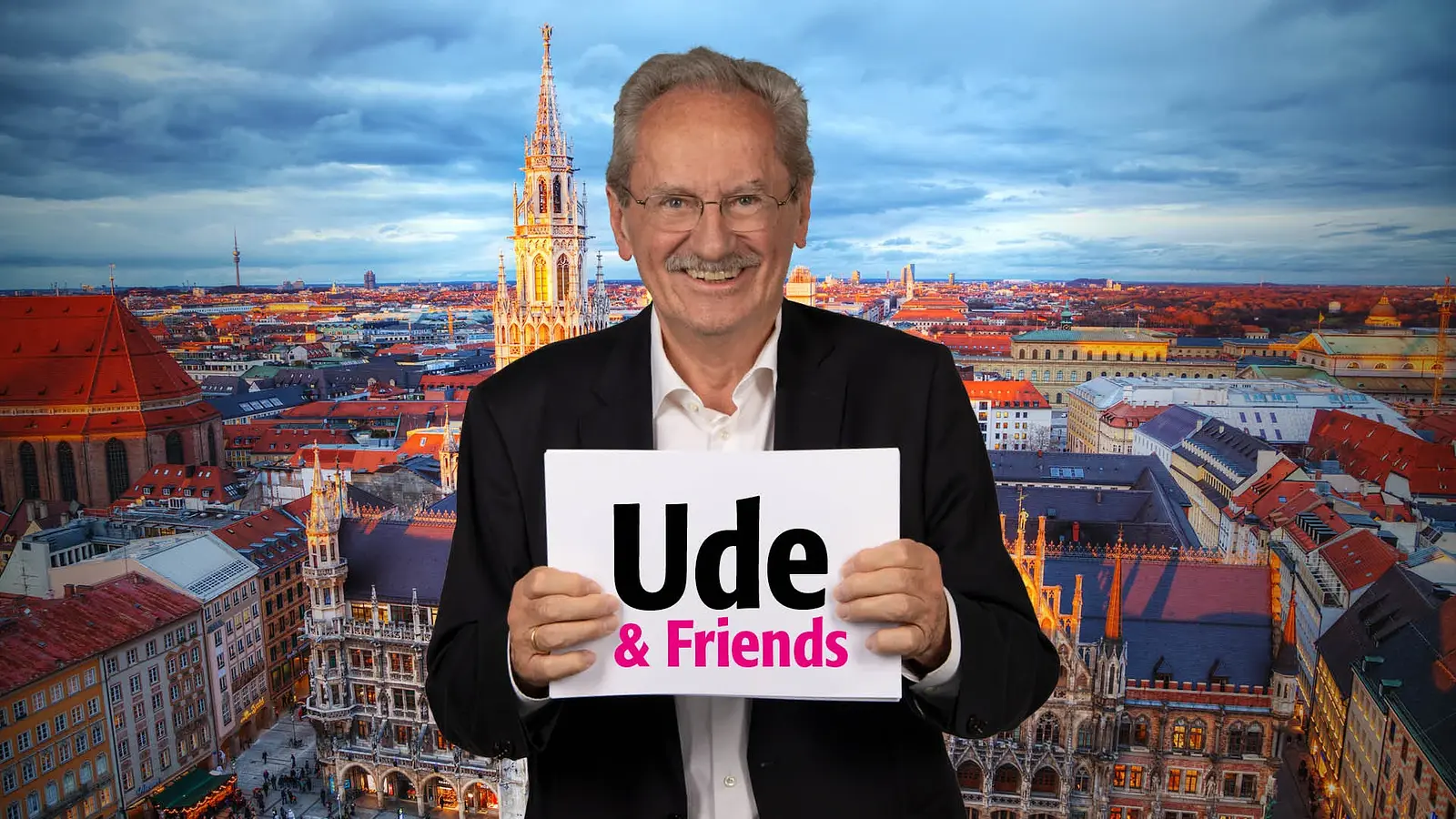 Person mit Brille hält ein Schild mit der Aufschrift Ude & Friends vor sich, im Hintergrund Panorama der Münchner Altstadt mit Rathaus, Kirchturm und Dächern bei Abendlicht